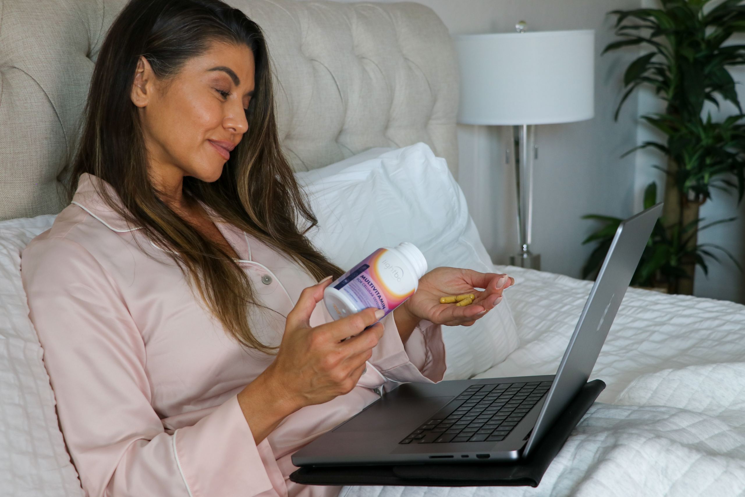 A scene featuring a woman comfortably seated in bed with a laptop on her lap, and in her hands, she holds a vial of dietary supplements for women.