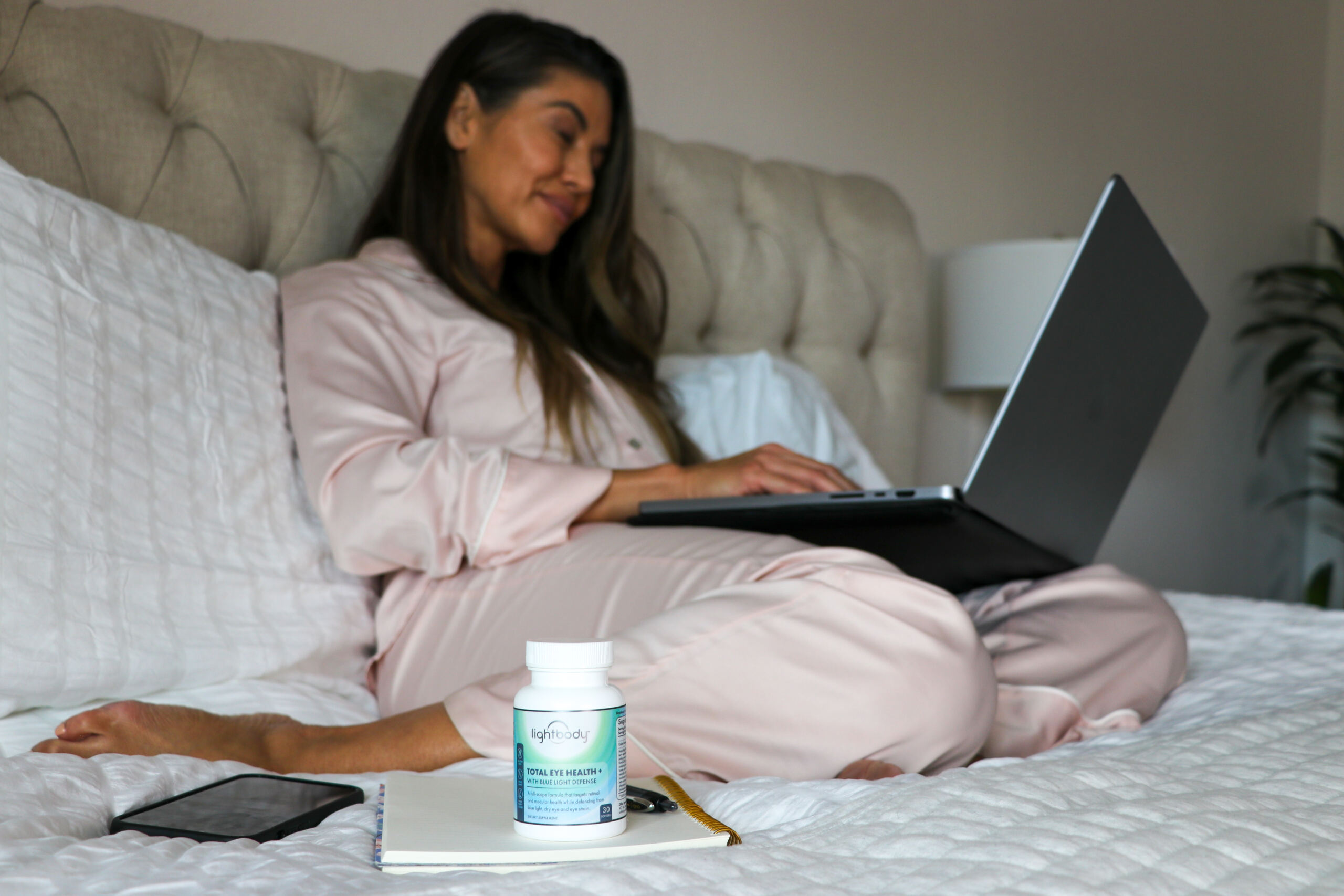 Woman sitting in bed, working on her laptop, with a bottle of supplements in the foreground - a photo illustrating a blog post trying to answer the question "Do eye health supplements work?"