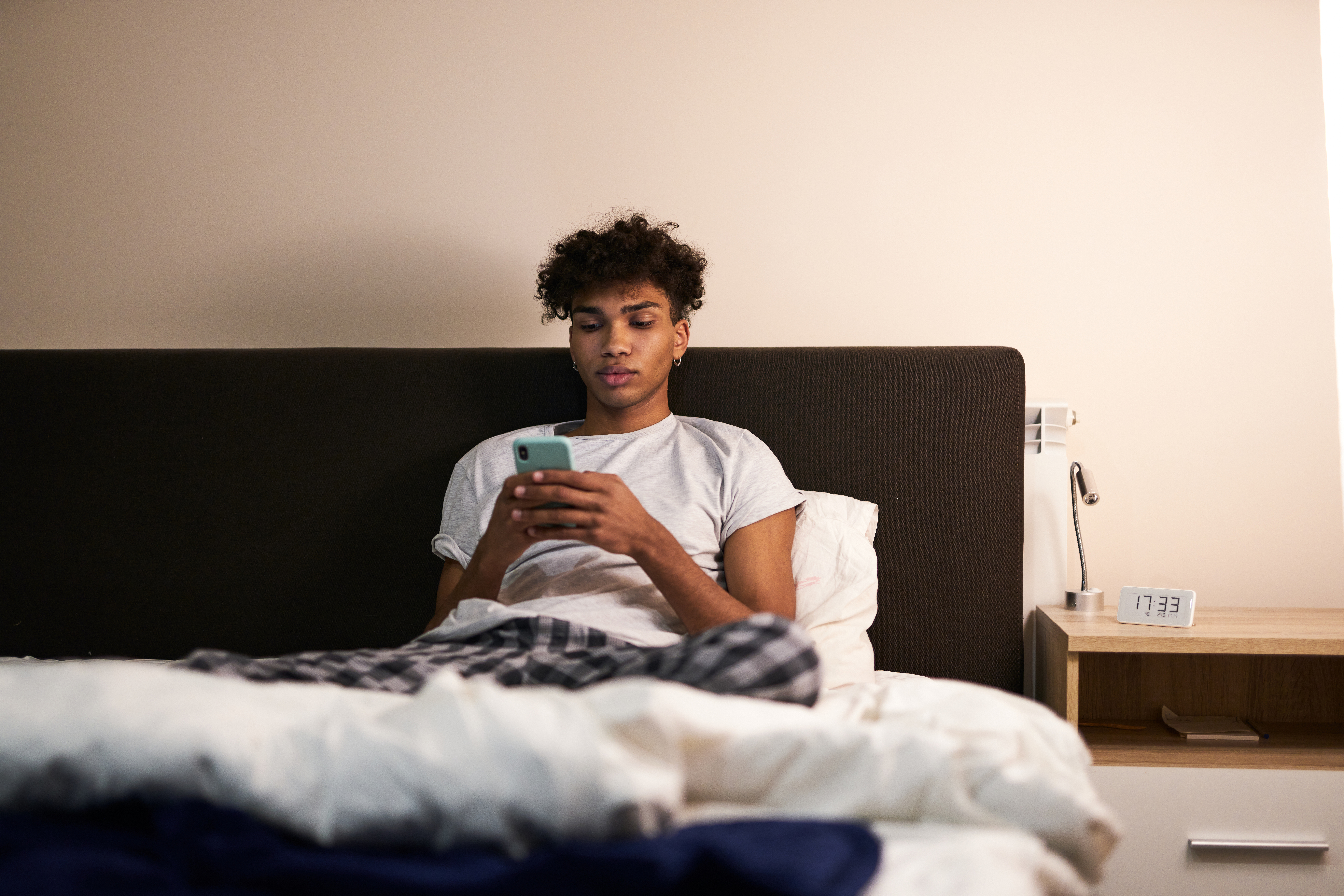 Young man lying in bed looking at his smartphone, illustrating a blog post about how does blue light affects your sleep.