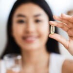 A young woman against a blurred background, holding an omega-3 pill in her hand in the foreground.